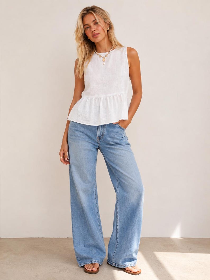 May include: A sleeveless white top with a round neckline and a ruffled peplum hem, paired with light blue denim jeans featuring a wide-leg cut and a classic five-pocket design. The outfit is completed with simple brown open-toe sandals.