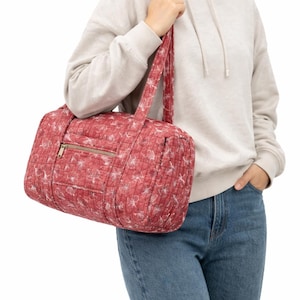 Pu&ograve; includere: Una borsa da viaggio trapuntata rossa con un motivo floreale bianco. La borsa ha una cerniera dorata e manici abbinati. La borsa viene portata a tracolla. &Egrave; una borsa da viaggio o da palestra.