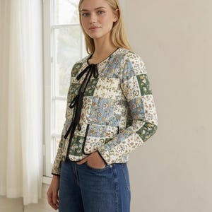 Może przedstawiać: Pikowana kurtka z patchworkowym wzorem z kwiatowymi wzorami w kolorze zielonym, niebieskim i kremowym. Kurtka ma czarne wykończenie i czarne wiązanie przy dekolcie. Rękawy są długie, a kurtka jest noszona z niebieskimi dżinsami.