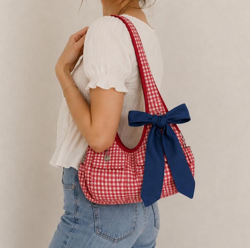 May include: A red and white gingham shoulder bag featuring a large navy blue fabric bow attached to the strap. The bag has a curved top opening, a single shoulder strap, and two front pockets with flaps. The fabric has a small silver charm.