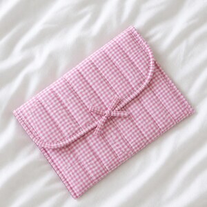 May include: A pink and white gingham patterned quilted clutch with a curved flap and a bow closure. The clutch is made of a soft, padded material and is lying on a white surface.