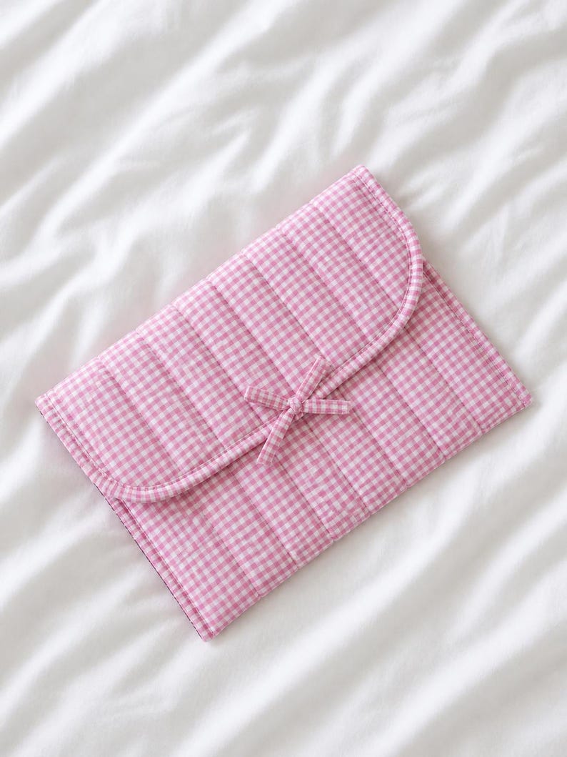 May include: A pink and white gingham patterned quilted clutch with a curved flap and a bow closure. The clutch is made of a soft, padded material and is lying on a white surface.