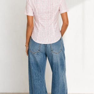 May include: A light pink short-sleeved shirt with a white and red plaid pattern, paired with blue denim wide-leg jeans. The shirt has a collared neckline and button-down front. The jeans have a high waist and a relaxed fit.