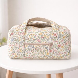 May include: A floral-patterned duffel bag with a cream background and a colorful array of small flowers. The bag has a beige zipper and handles. The bag is sitting on a white table.
