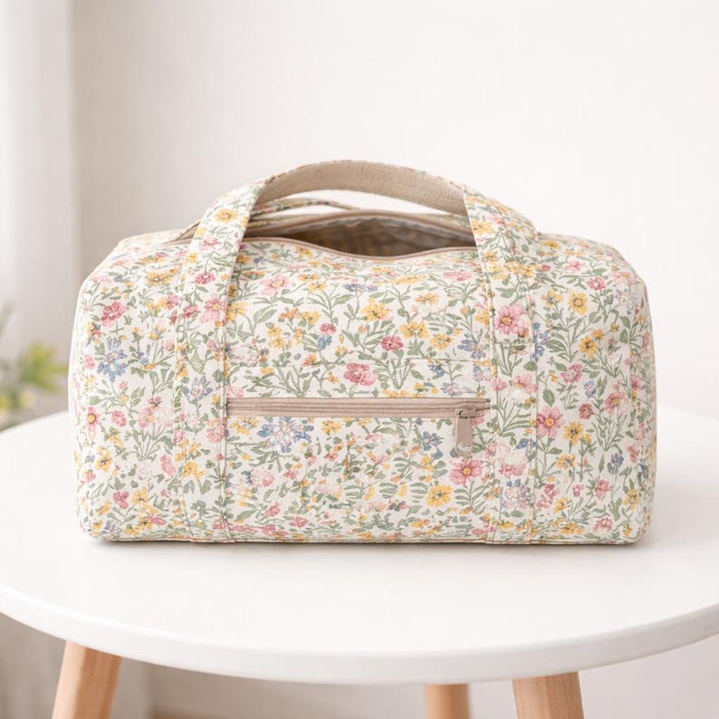 May include: A floral-patterned duffel bag with a cream background and a colorful array of small flowers. The bag has a beige zipper and handles. The bag is sitting on a white table.