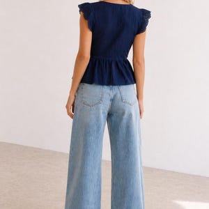 May include: A woman wearing a navy blue top with eyelet detailing on the sleeves and a peplum waist, paired with light blue wide-leg jeans. The outfit is casual and stylish.