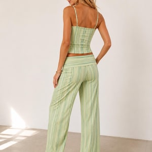 May include: A two-piece outfit featuring a striped camisole top and matching wide-leg pants. The set is in shades of green and white, with vertical stripes. The top has thin straps, and the pants have a high waist. The outfit is made of a lightweight fabric.