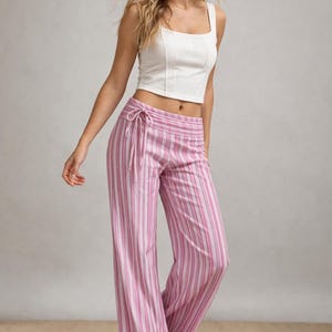 May include: A woman wearing a white crop top and pink and white striped palazzo pants. The pants have a drawstring waist. The outfit is casual and summery.