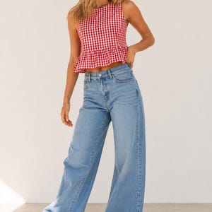 May include: A sleeveless red and white gingham peplum top paired with light blue wide-leg denim jeans and white sneakers. The top features a round neckline and a ruffled hem, while the jeans have a classic five-pocket design and a button fly.