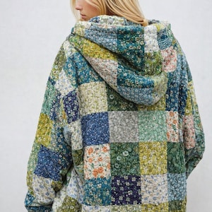 May include: A quilted hoodie jacket with a patchwork design. The jacket features a variety of floral patterns in shades of blue, green, and orange. The jacket has a hood and a relaxed fit, perfect for layering.