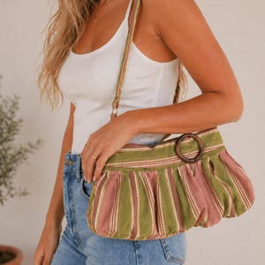 May include: A green and pink striped handbag with a wooden handle. The bag has a pleated design and a long strap. The bag is being held by a person wearing a white tank top and blue jeans.