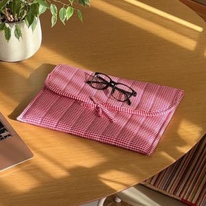May include: A red and white gingham patterned glasses case with a black frame pair of glasses on top. The case is closed with a matching fabric tie. The case is on a wooden table.
