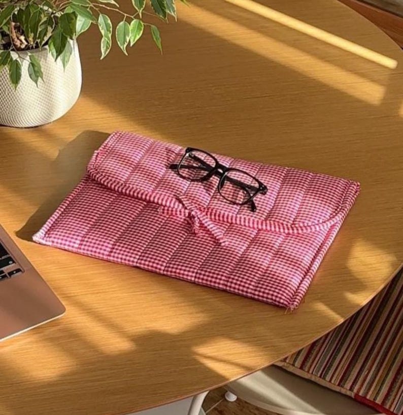 May include: A red and white gingham patterned glasses case with a black frame pair of glasses on top. The case is closed with a matching fabric tie. The case is on a wooden table.