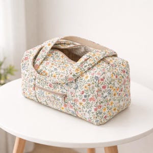 May include: A floral print duffel bag with a cream background and colorful flowers in pink, yellow, and blue. The bag has a zippered top and a front zippered pocket. It features two handles and is sitting on a white table.