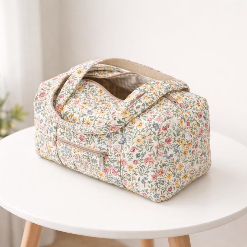 May include: A floral print duffel bag with a cream background and colorful flowers in pink, yellow, and blue. The bag has a zippered top and a front zippered pocket. It features two handles and is sitting on a white table.