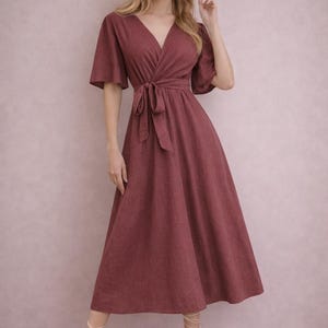 Op de afbeelding: Een bordeauxrode wikkeljurk met korte mouwen en een strik in de taille. De jurk heeft een V-hals en een uitlopende rok. Het model draagt witte sandalen met bandjes. De jurk is gemaakt van een textuurstof.