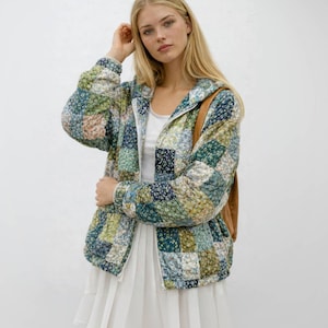 May include: A patchwork-style hooded jacket in shades of blue, green, and cream. The jacket features a floral pattern and a zipper closure. The model is wearing a white top and skirt, and a brown bag is over their shoulder.