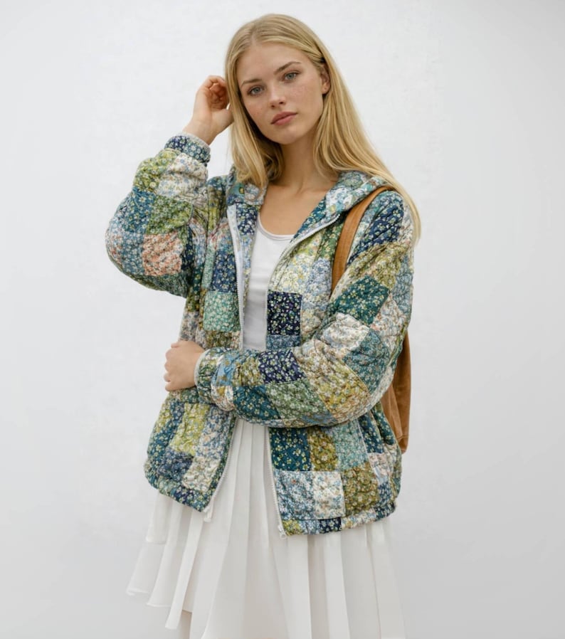 May include: A patchwork-style hooded jacket in shades of blue, green, and cream. The jacket features a floral pattern and a zipper closure. The model is wearing a white top and skirt, and a brown bag is over their shoulder.