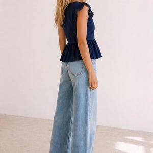 May include: A woman wearing a navy blue top with a peplum waist and eyelet detailing on the sleeves, paired with light wash wide-leg jeans. The jeans have a high waist and a relaxed fit.