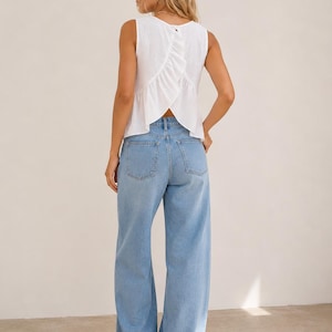 May include: A sleeveless white top with a textured fabric and a vertical ruffle detail along the back, paired with light blue denim jeans featuring a wide-leg cut, a high waist, and two back pockets. The top has a button closure at the neckline.