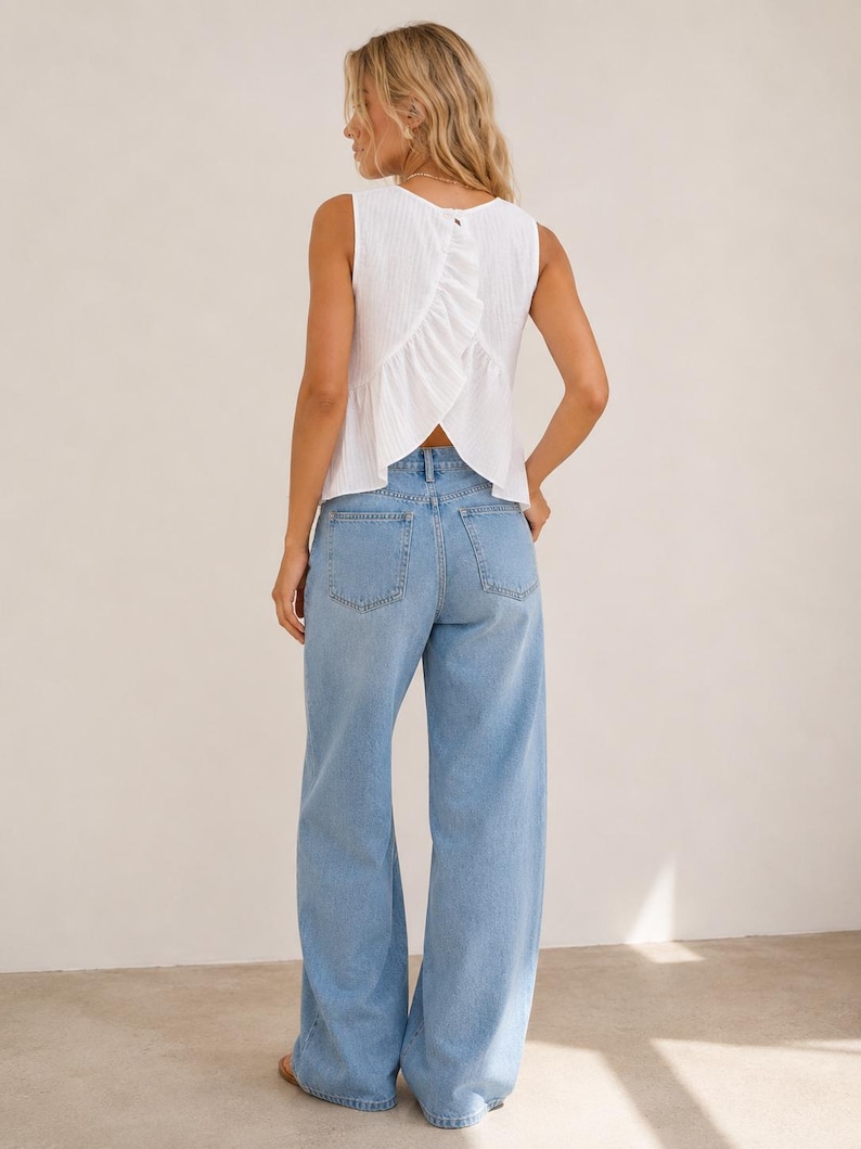May include: A sleeveless white top with a textured fabric and a vertical ruffle detail along the back, paired with light blue denim jeans featuring a wide-leg cut, a high waist, and two back pockets. The top has a button closure at the neckline.