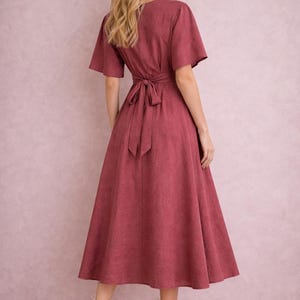 Op de afbeelding: Een midi-jurk in een stoffige roze tint, met korte mouwen en een strik in de taille. De jurk heeft een uitlopende rok en is gemaakt van een textuurstof. Het model draagt witte sandalen met bandjes. De jurk is ongeveer 110 cm lang.