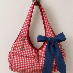 May include: A red and white gingham shoulder bag featuring a large navy blue fabric bow attached to the front. The bag has two exterior pockets, a red interior lining, and a silver-toned metal heart charm hanging from a small ring on the strap.