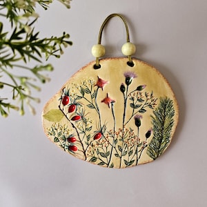 Op de afbeelding: Een keramische wandhanger met een botanisch ontwerp. Het stuk is lichtbeige van kleur met een handgeschilderde illustratie van wilde bloemen, bessen en groen. Het heeft een leren lus om op te hangen en twee houten kralen aan de bovenkant.