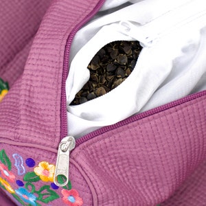 K&ouml;nnte beinhalten: Eine violette gesteppte Tasche mit gestickten Blumen und einem Rei&szlig;verschluss, der sich &ouml;ffnet und einen wei&szlig;en Beutel mit Buchweizenh&uuml;lsen enth&uuml;llt.