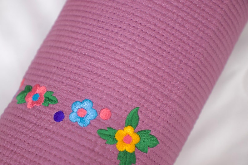 K&ouml;nnte beinhalten: Ein lila gestepptes Kissen mit gestickten Blumen in den Farben Rosa, Blau, Gelb und Gr&uuml;n.