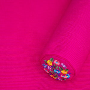 K&ouml;nnte beinhalten: Ein rosa gestepptes Kissen mit gestickten Blumendesigns in verschiedenen Farben. Das Kissen ist aufgerollt und hat eine runde Form.