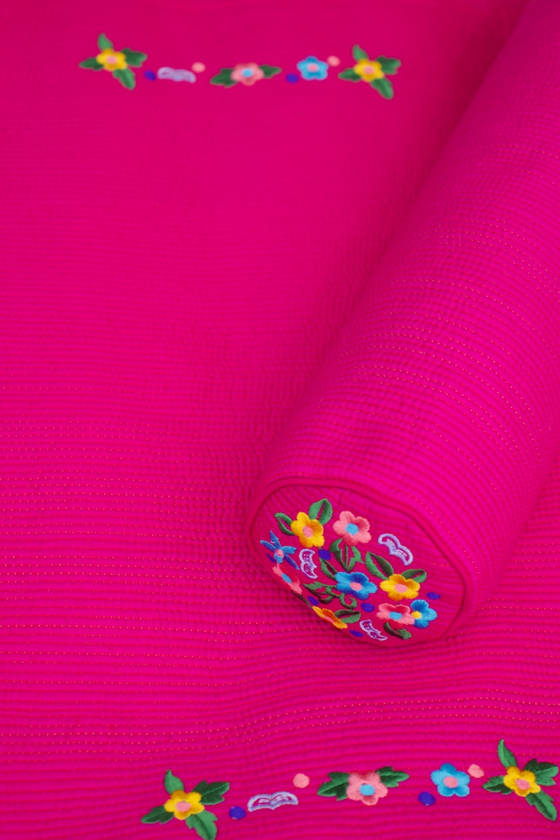 K&ouml;nnte beinhalten: Ein rosa gestepptes Kissen mit gestickten Blumendesigns in verschiedenen Farben. Das Kissen ist aufgerollt und hat eine runde Form.