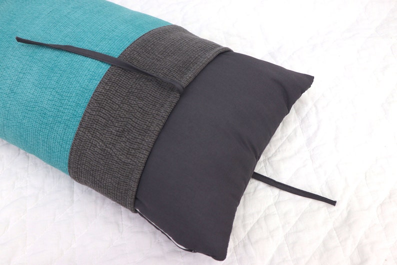 May include: A long teal and gray pillow with a black tie closure. The pillow is lying on a white surface.