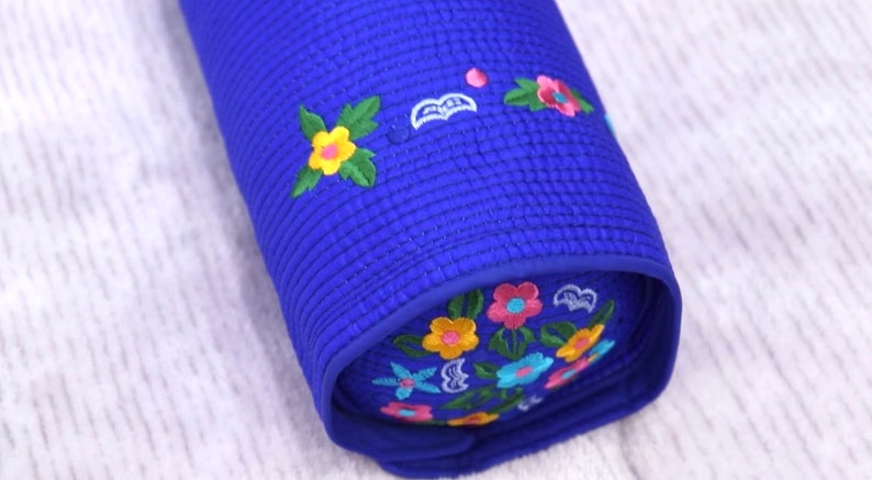 K&ouml;nnte beinhalten: Ein blaues, zylindrisches Kissen mit gestickten Blumen in Gelb-, Rosa-, Blau- und Gr&uuml;nt&ouml;nen.