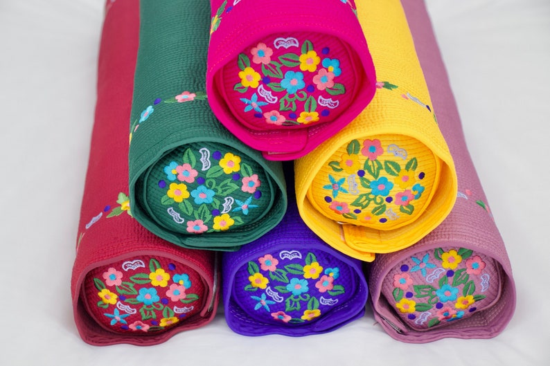 K&ouml;nnte beinhalten: Sechs bunte zylindrische Kissen mit gestickten Blumenmustern. Die Kissen sind pyramidenf&ouml;rmig gestapelt, wobei das oberste Kissen rosa ist, gefolgt von gelb, gr&uuml;n, rot, lila und schlie&szlig;lich einem helllila Kissen unten.