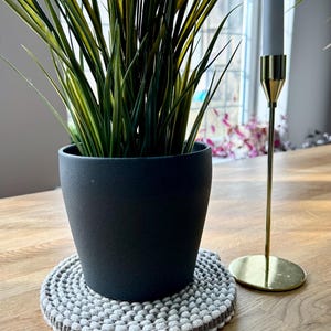 May include: A black pot with a green plant sits on a round, white and grey crocheted coaster. A gold candle holder with a white candle is on the right side of the image.