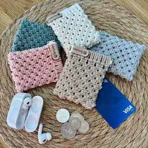 May include: Five crocheted coin purses in a variety of colours, including pink, beige, white, and grey. Each purse has a small wooden tag with the text "Nicky Krafty" on it. A blue VISA debit card and a set of white wireless earbuds are also in the image.