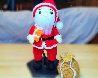 Puede incluir: Un muñeco de Papá Noel de crochet que lleva un traje rojo con ribetes blancos y una barba blanca. Lleva un pequeño regalo naranja y está de pie sobre una base negra. Un saco de crochet marrón está a su lado.