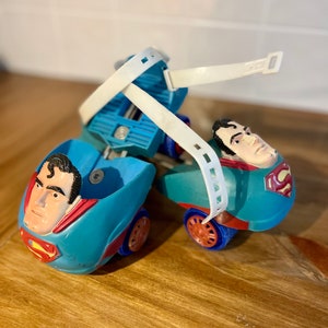 May include: A pair of blue and red roller skates with Superman's face on the front. The skates have white straps and wheels.