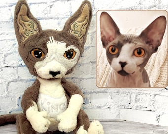 May include: A plush toy modeled after a Sphynx cat with brown and white fur, large pointed ears, and amber-colored eyes. The toy features a white chest patch with the embroidered word KING and is displayed next to a photograph of the real cat.
