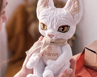 May include: A plush white cat toy with large pointed ears, textured fur, and amber-colored eyes. The cat wears a beige scarf and a matching chest patch embroidered with the name Baron in brown thread. The toy has a soft, stuffed appearance.