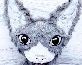 May include: A plush toy shaped like a cat head with long, pointed ears. The toy is covered in gray faux fur with a white muzzle and nose area. It features large, dark brown embroidered eyes and intricate light gray stitching details on the ears.