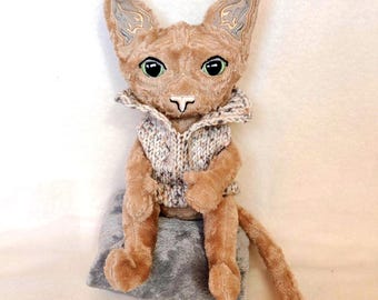May include: A plush toy resembling a Sphynx cat with tan fur, large pointed ears, and green eyes. The cat wears a beige and grey speckled knit vest with a collar. It is seated on a small, rectangular grey plush cushion with a long tail extending out.