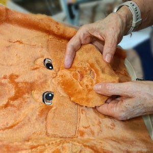 May include: A soft orange plush fabric piece featuring embroidered details of two dark eyes with white highlights and a separate, detached orange plush muzzle section with an embroidered nose, showcasing the texture of the fuzzy material.