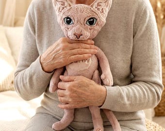 May include: A plush, pink Sphynx cat doll with large blue eyes and a heart-shaped patch on its chest. The doll is held by a person wearing a gray sweater. The doll's ears are large and pointed.