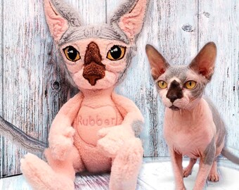May include: A plush toy resembling a Sphynx cat with light pink fur, large pointed ears, and a dark brown nose, featuring a heart-shaped patch on its chest with the word Rubber embroidered in pink thread, positioned next to a real Sphynx cat.