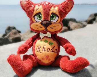 May include: A plush cat toy with deep red fur, large green eyes, and a light orange face patch. The chest features a light orange heart-shaped panel embroidered with the name Rhoda, three small green flowers, and three small orange hearts.