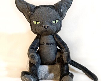 May include: A plush toy shaped like a dark gray cat with large pointed ears, green eyes, and a stern expression. The chest area features the embroidered text David Meowie. The figure has a textured, fabric-covered body with a long tail and paws.