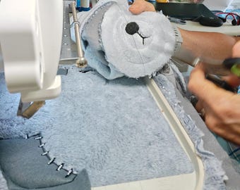 May include: A light blue plush fabric piece with a dark gray stitched nose and mouth detail, secured in a white embroidery hoop under a sewing machine needle. The fabric features a dark gray patch attached with visible black thread stitches.