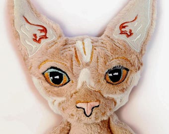 May include: A plush toy shaped like a Sphynx cat head with light tan fur, large pointed ears featuring embroidered red accents, and wide amber-colored eyes. The face has detailed stitching, a pink nose, and a soft, textured fabric construction.